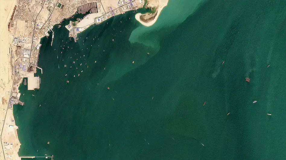 Satellite image showing a coastal city with buildings, sandy land, harbor docks, and numerous small boats scattered across the greenish-blue water. A small sandy peninsula extends into the sea near the center-top.