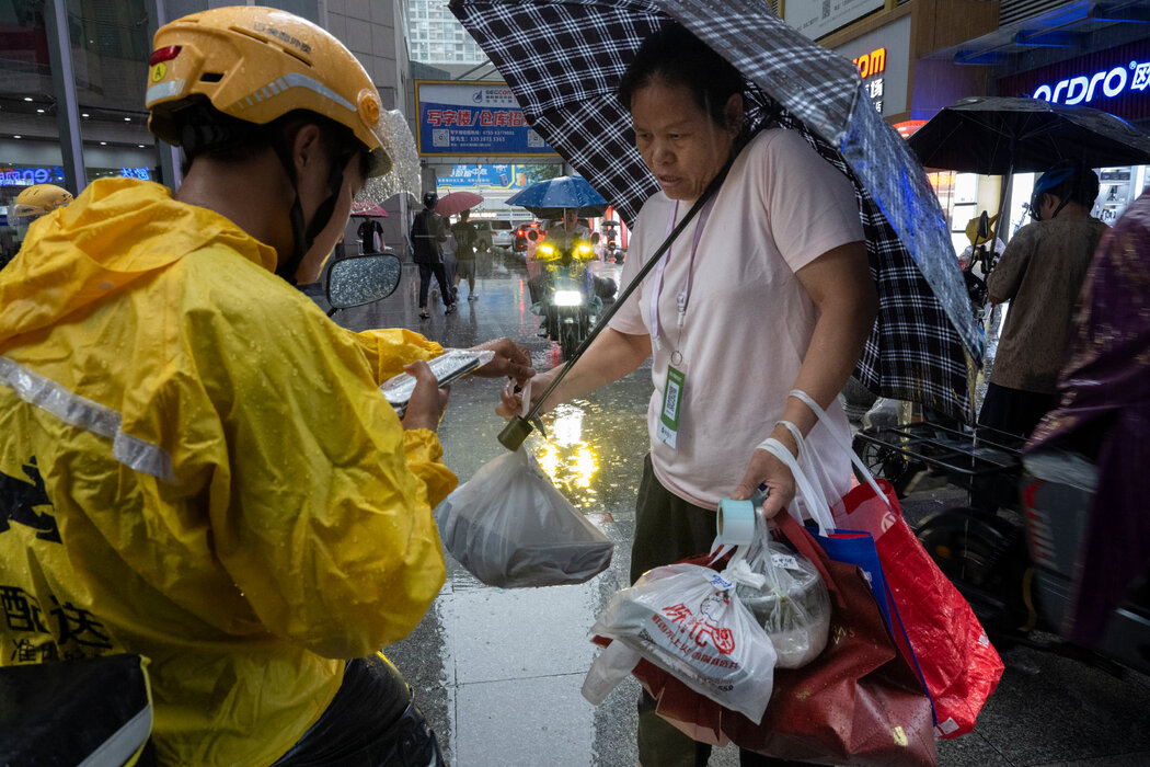 在赛格广场外,一名骑电动车的外卖骑手将餐食交给一位代送跑腿员,由他送到大楼里下单的顾客手中。